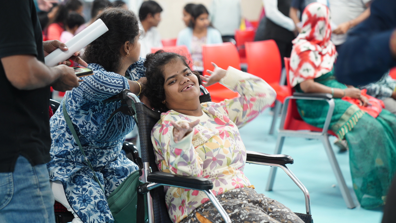 A smiling young woman in a wheelchair waving at a DigiAbility community camp, with other attendees seated around her